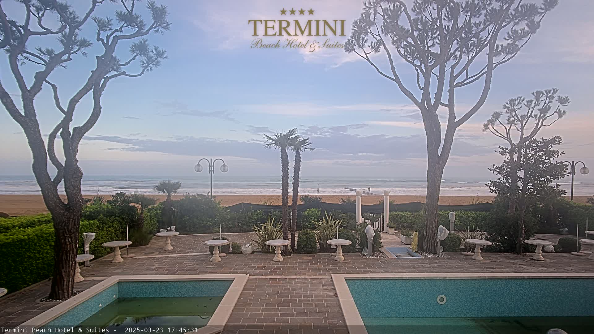 A patio with two pools overlooks a calm ocean beach under a mostly clear, pale sky.