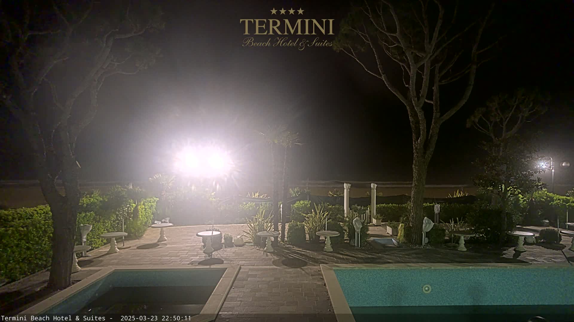 A nighttime view of a swimming pool and patio area, surrounded by trees and shrubs, with hazy or foggy weather conditions.