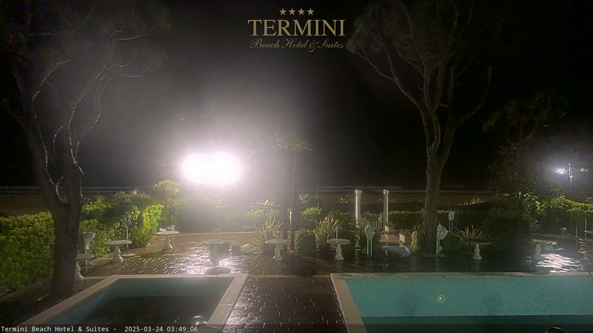 At night, under a dark, possibly stormy sky, a wet patio area with empty tables and two pools is visible, next to a beach.