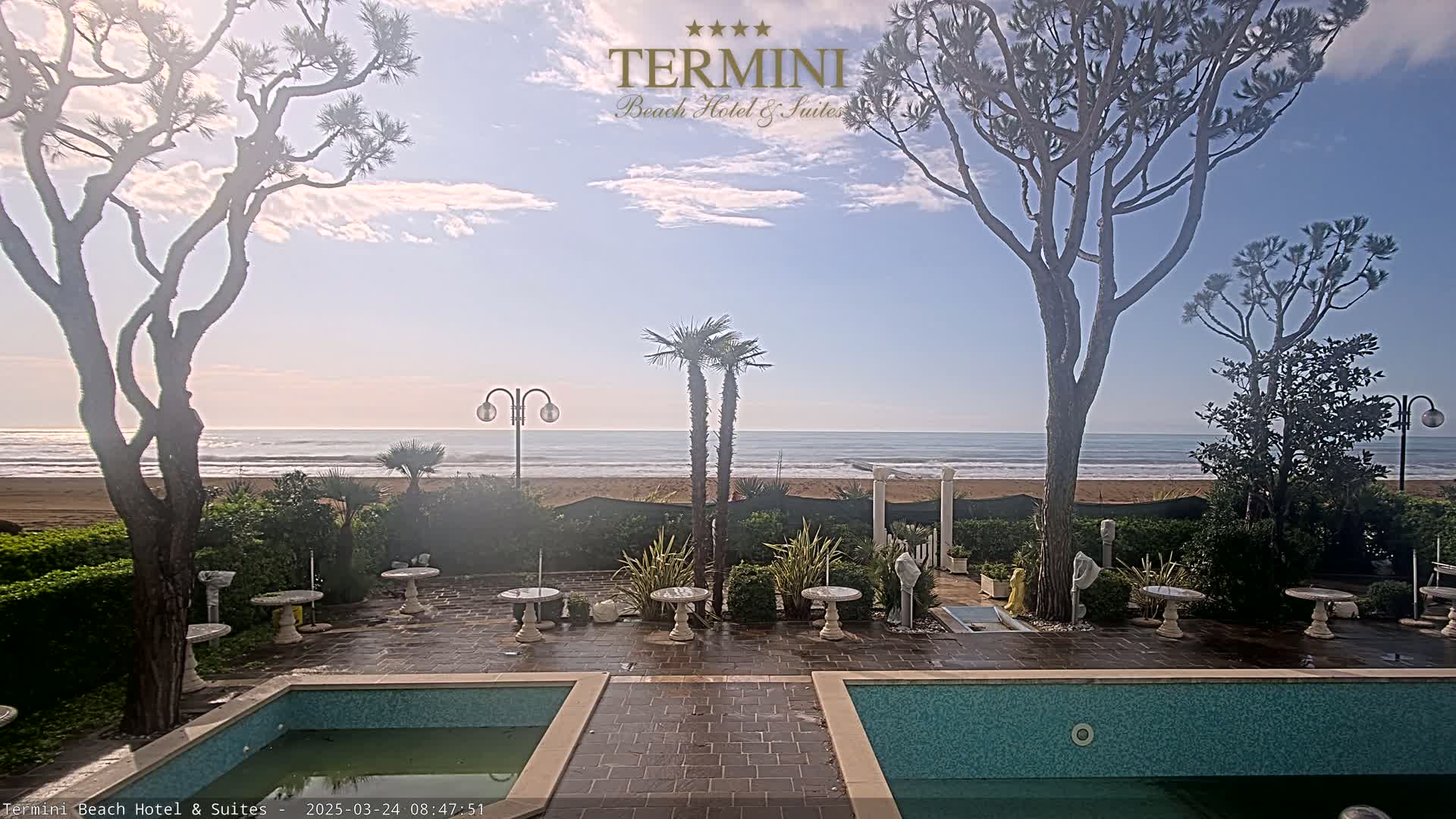 A tranquil beach scene is visible from a patio with two pools, featuring palm trees, stone tables, and a calm ocean under a partly cloudy sky.
