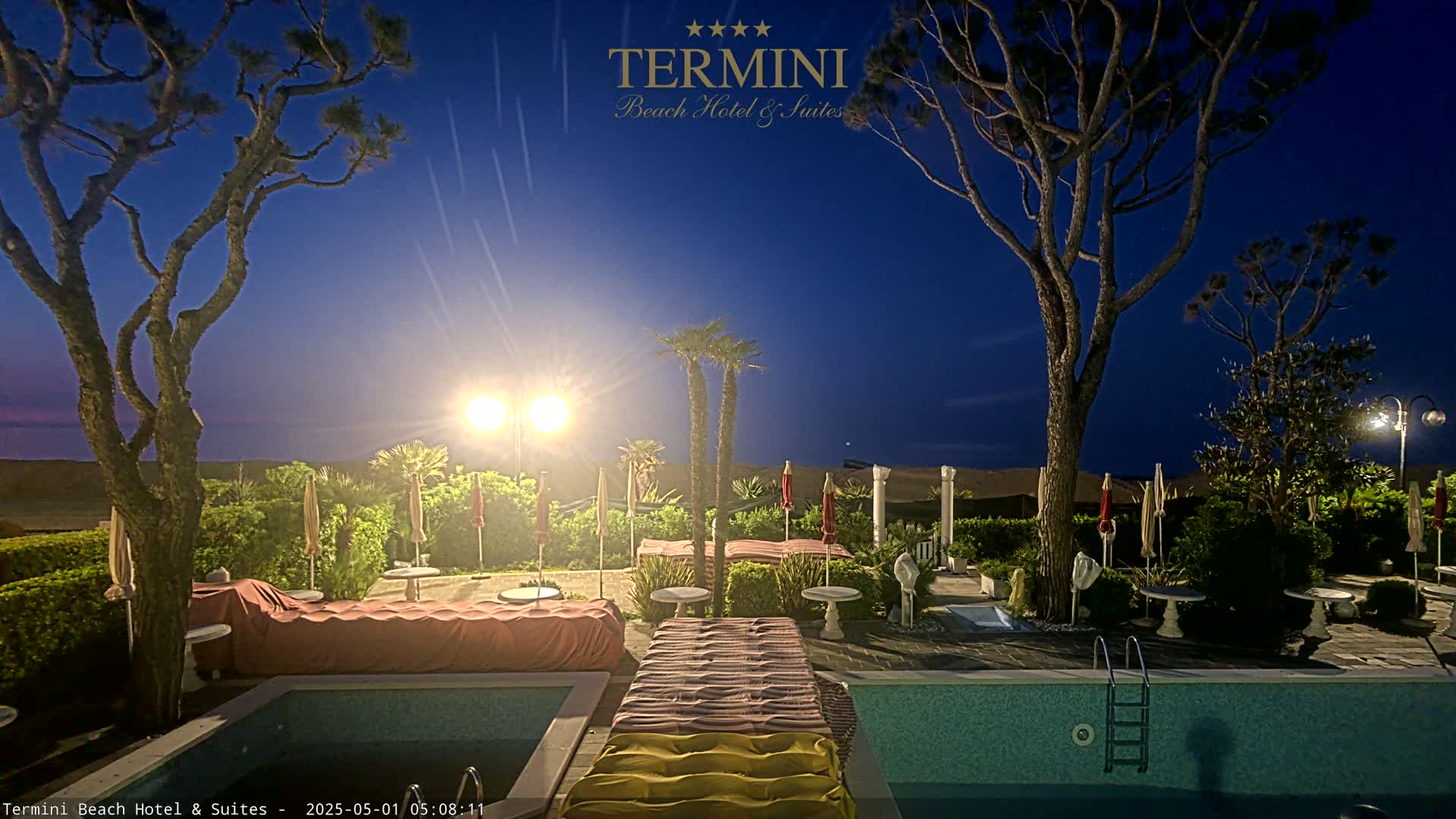 A nighttime scene shows a swimming pool area with lounge chairs, umbrellas, and trees illuminated under a clear, dark blue sky.