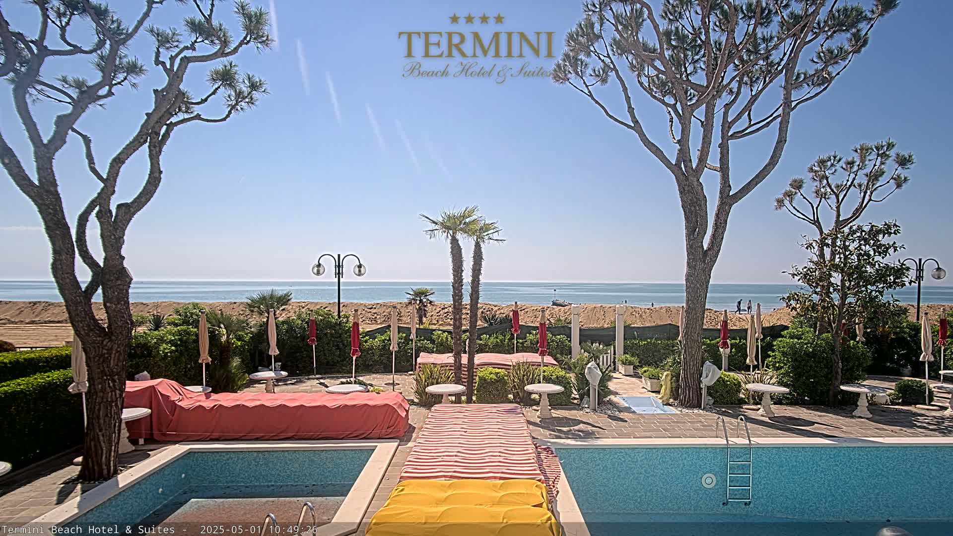 A swimming pool and patio area overlook a sandy beach and calm ocean under a clear blue sky.