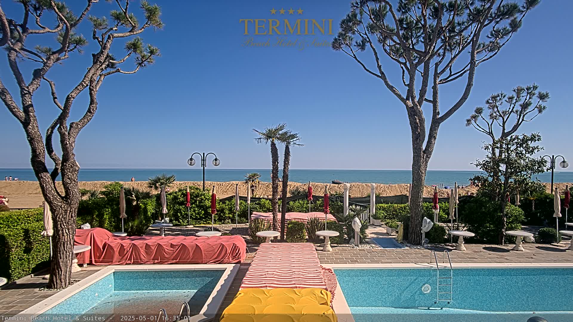 A swimming pool and patio area are situated beside a sandy beach and calm ocean under a clear blue sky.