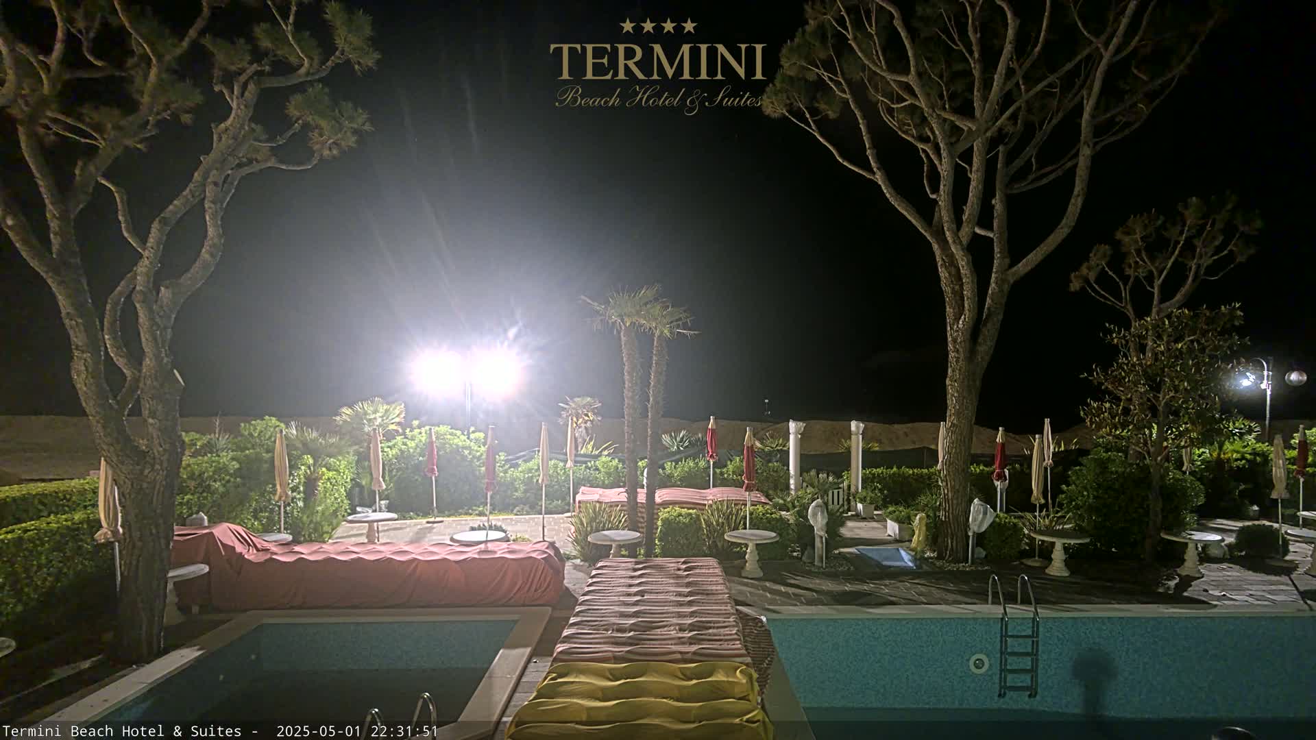 A nighttime view of a swimming pool area at a resort, illuminated by bright lights, with trees, closed umbrellas, and lounge chairs surrounding the pool.