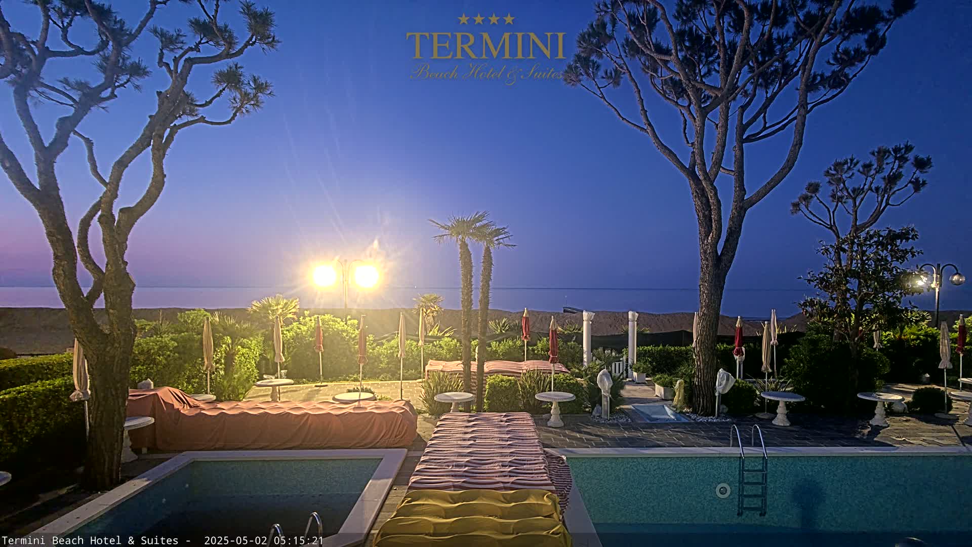 A twilight view of a hotel pool area, featuring palm trees, beach umbrellas, and a calm sea in the background.