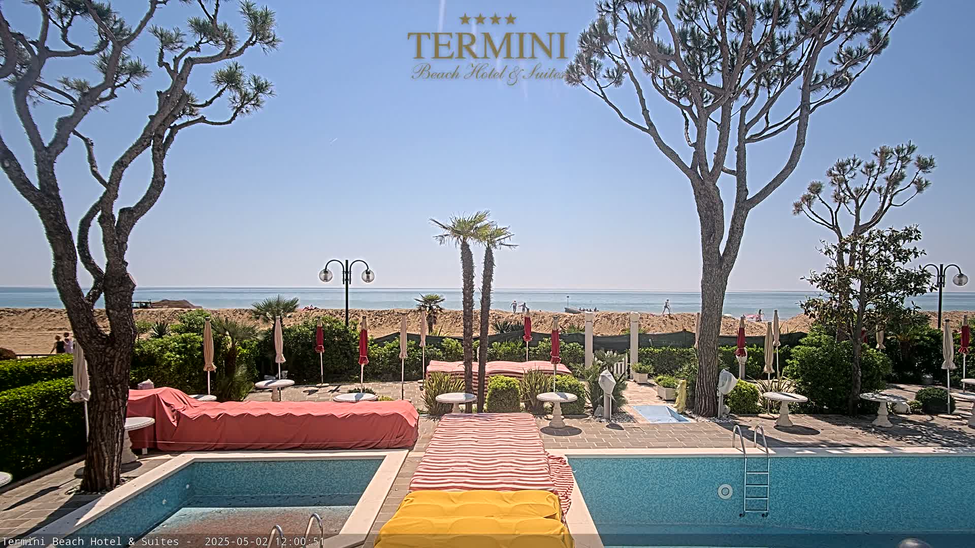 A resort pool area with lounge chairs and umbrellas overlooks a sandy beach and calm ocean under a clear blue sky.