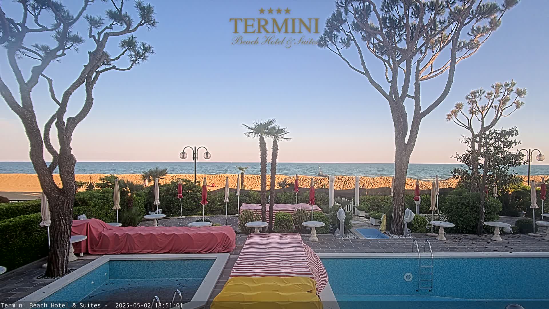 A swimming pool and patio area overlook a calm ocean under a clear, light-blue sky.