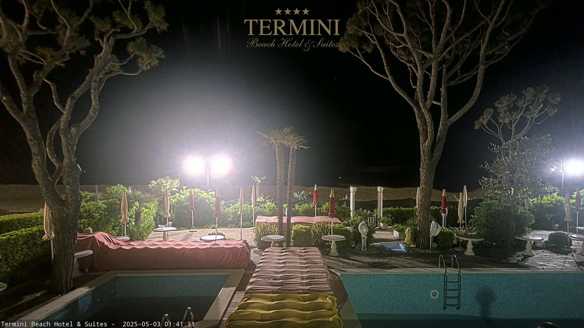 A nighttime view of a swimming pool area with closed umbrellas, lounge chairs covered, and tall trees illuminated by lights, near a sandy beach.