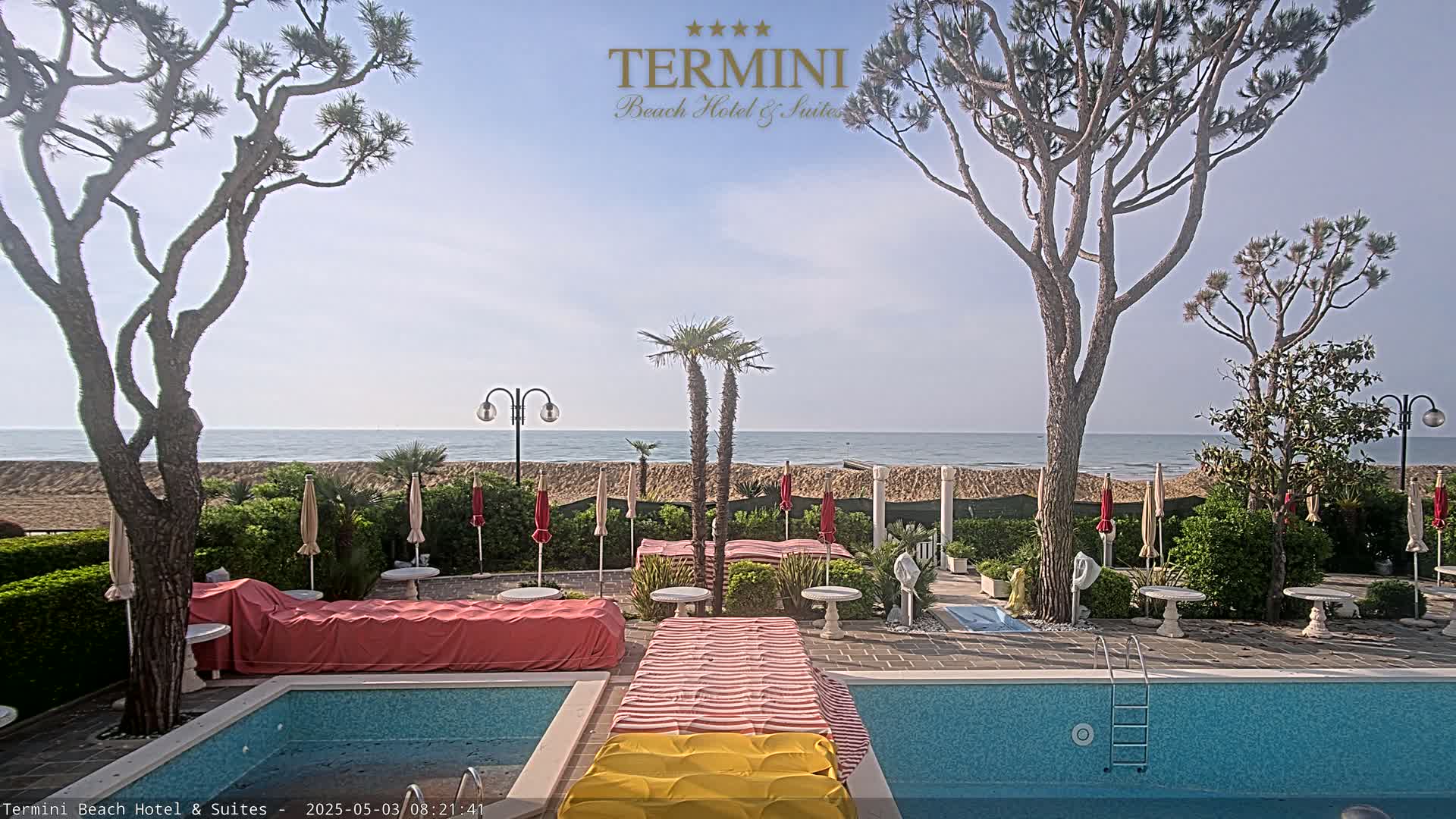 A swimming pool and patio area, partially covered with fabric, overlook a calm ocean beach under a clear sky.