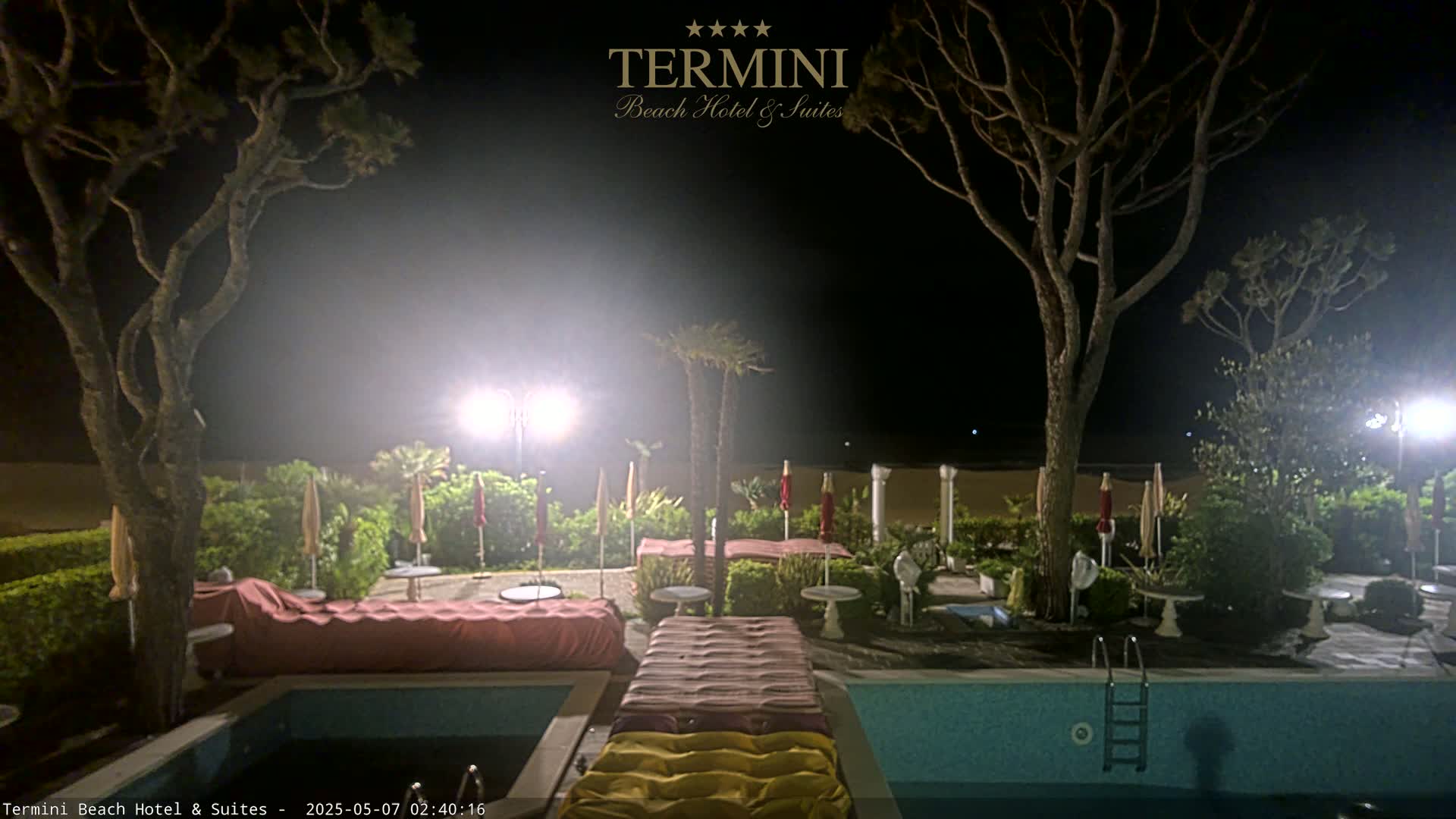 A nighttime view of a resort pool area with beach access, illuminated by lights, featuring several trees, lounge chairs, and umbrellas around the pool.