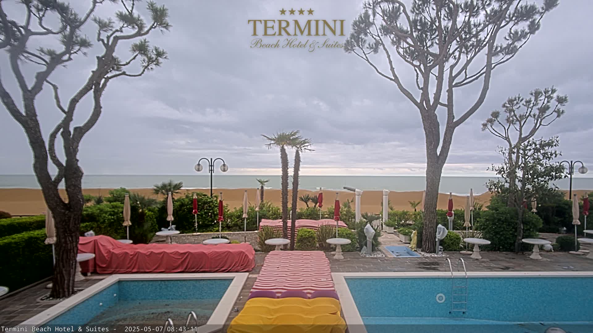 A swimming pool and patio area overlook a sandy beach and calm ocean under an overcast sky.