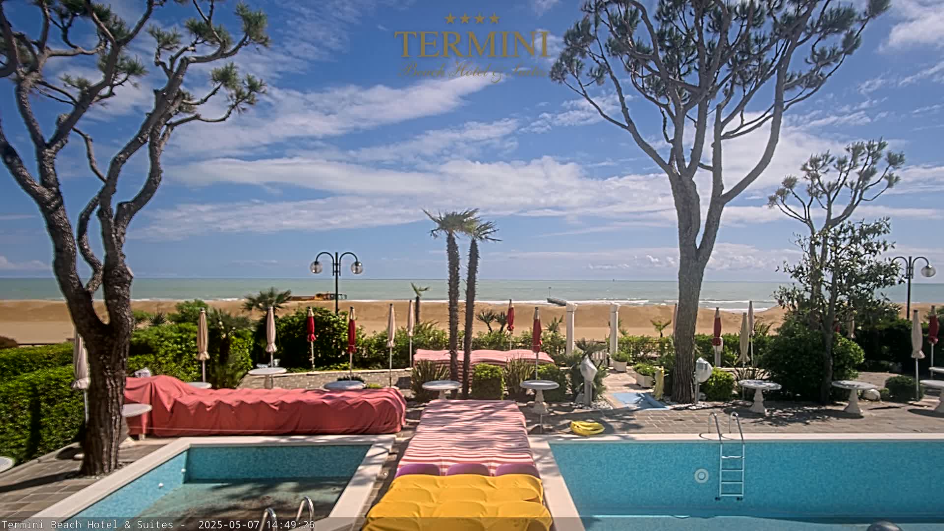 A resort pool area with lounge chairs and umbrellas overlooks a sandy beach and calm ocean under a partly cloudy sky.