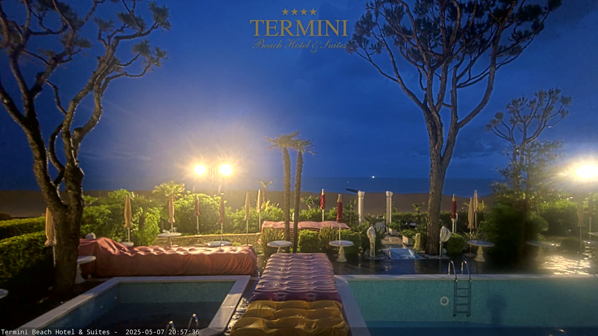 A twilight scene shows a swimming pool with inflatable loungers next to a landscaped area with palm trees and umbrellas, overlooking a calm ocean.