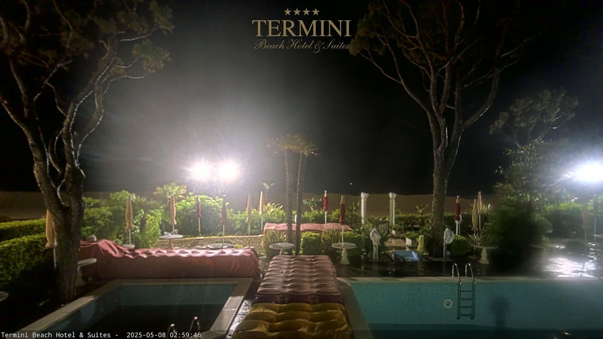 A nighttime view of a pool area with outdoor lights, trees, and closed umbrellas surrounding a swimming pool with air mattresses near the water's edge, under foggy conditions.