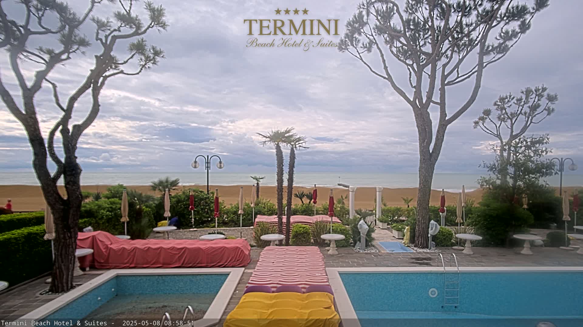 A resort's pool area with two pools overlooks a sandy beach and calm ocean under a cloudy sky.