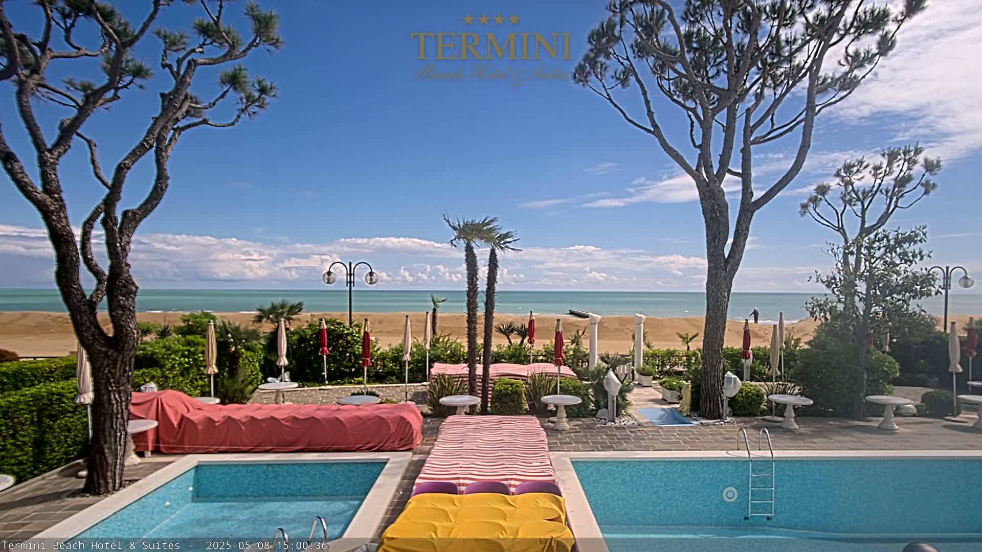 A swimming pool area with lounge chairs and umbrellas sits between two trees overlooking a sandy beach and calm ocean under a partly cloudy sky.