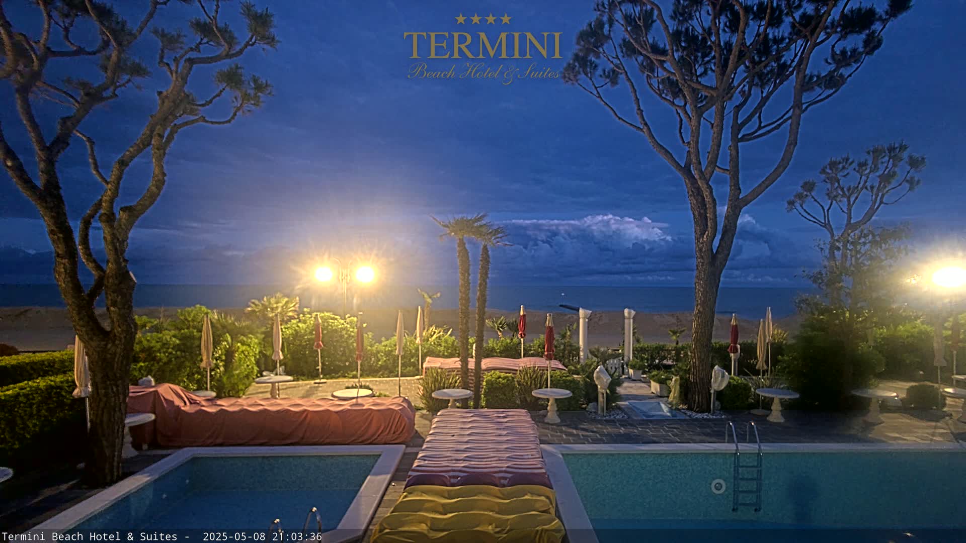 A nighttime view of a resort pool area with a beach and ocean in the background under a twilight sky.