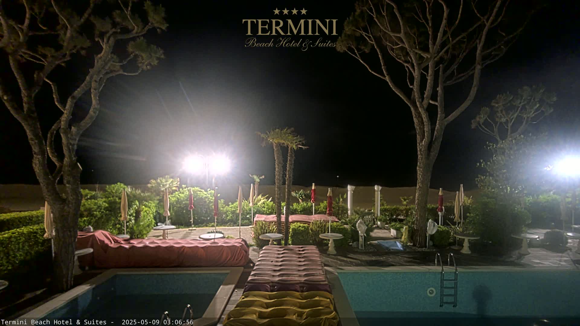 At night, a swimming pool with lounge chairs and umbrellas sits beside a sandy beach illuminated by bright lights, surrounded by trees and landscaping.