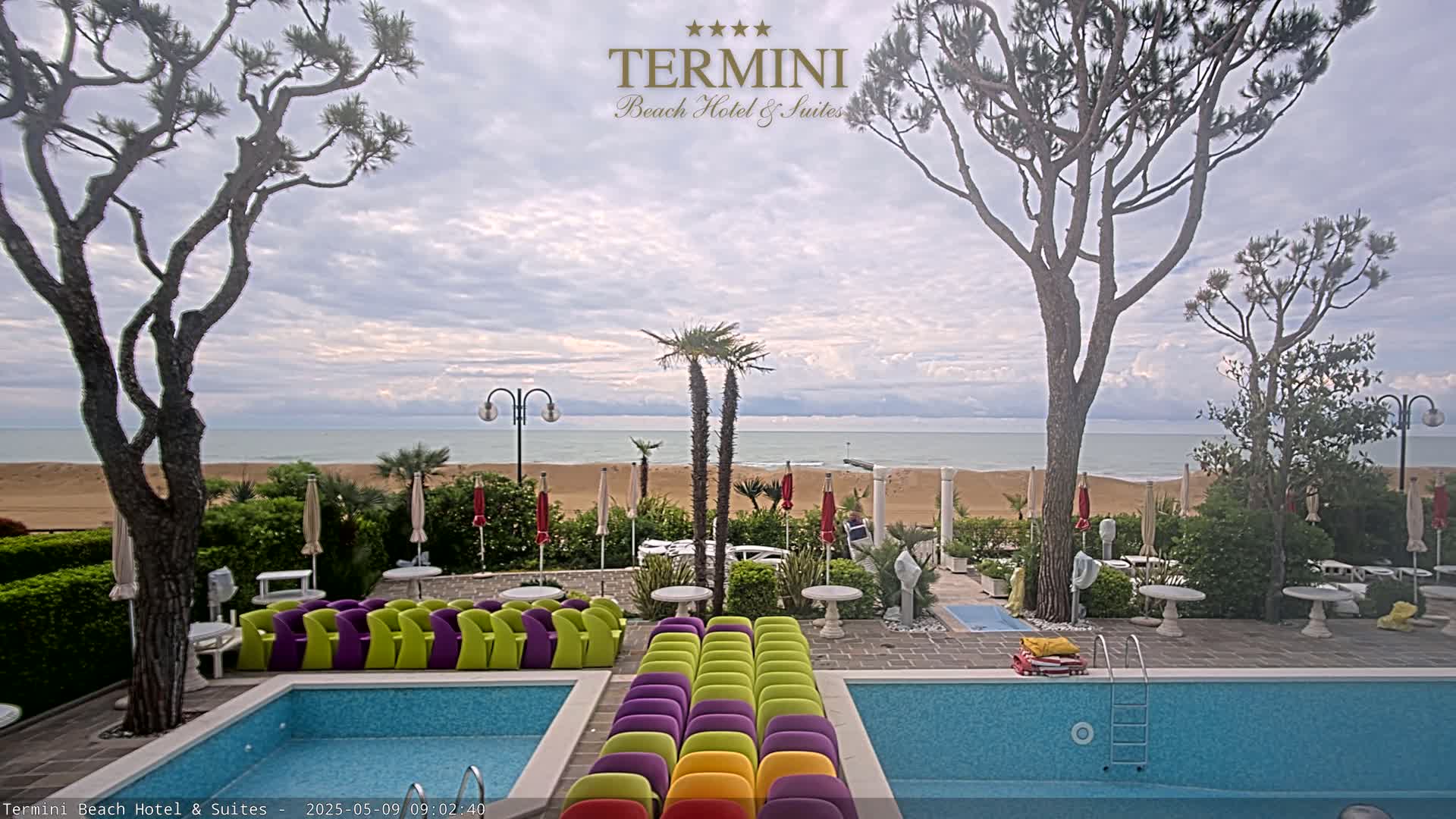 A hotel pool area with colorful chairs sits between two pools, overlooking a sandy beach and calm ocean under a partly cloudy sky.