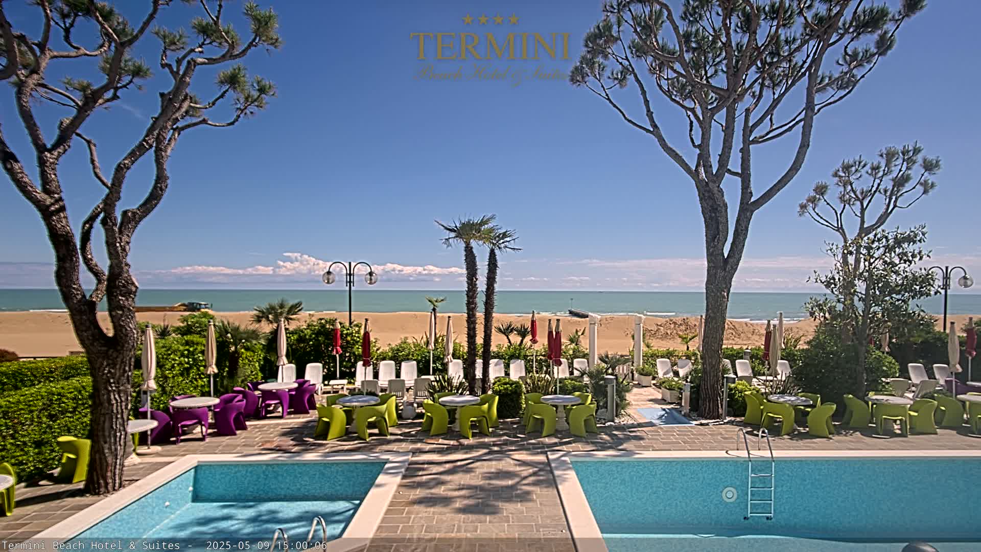 A beachfront hotel pool area with colorful patio furniture, palm trees, and a clear blue sky overlooks a sandy beach and calm ocean.
