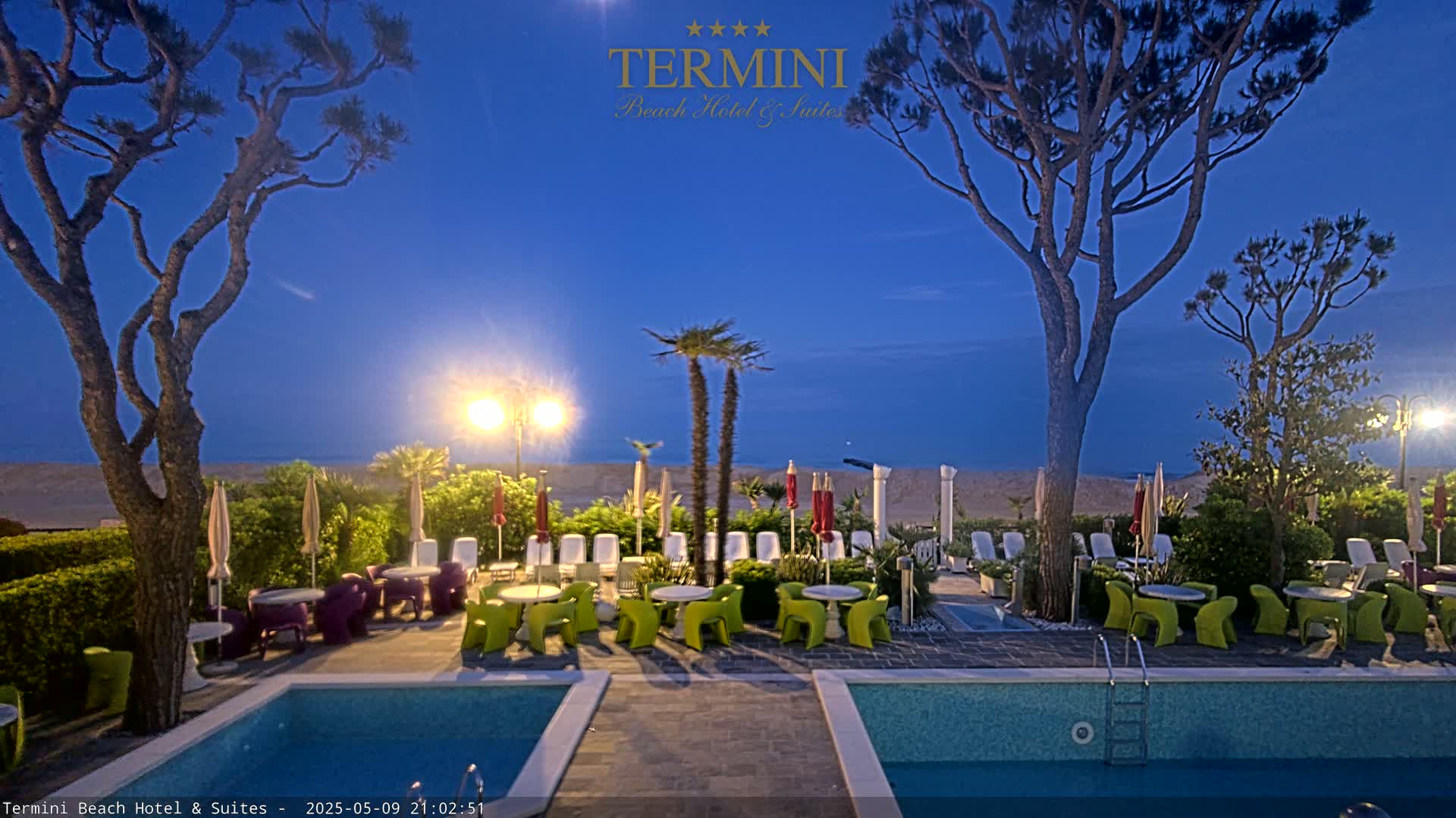 Under a twilight sky, a hotel pool area features two pools, outdoor furniture, and palm trees near a sandy beach.