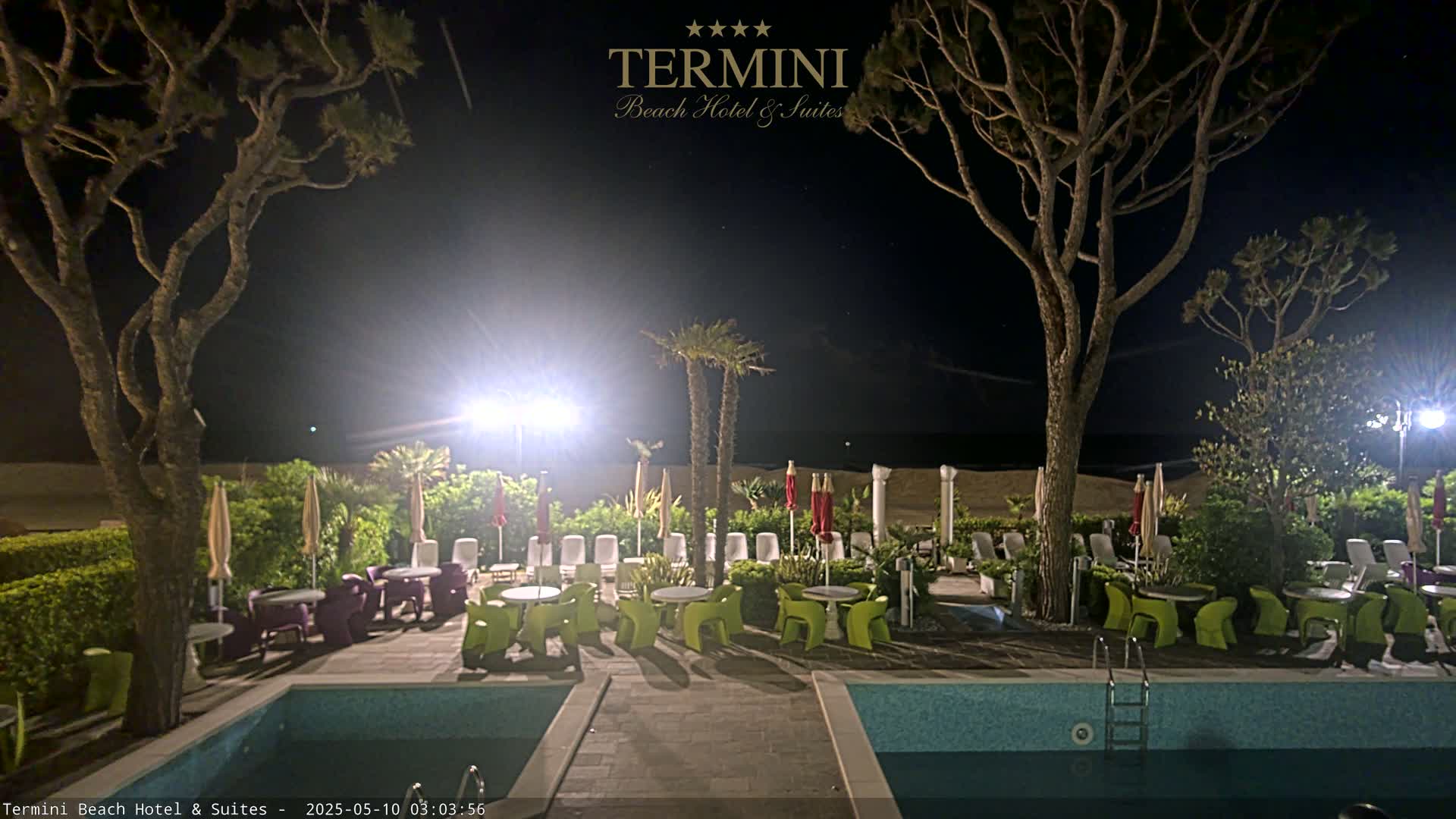 A nighttime view of a resort pool area with lounge chairs, tables, and palm trees surrounding a swimming pool under a clear, dark sky.
