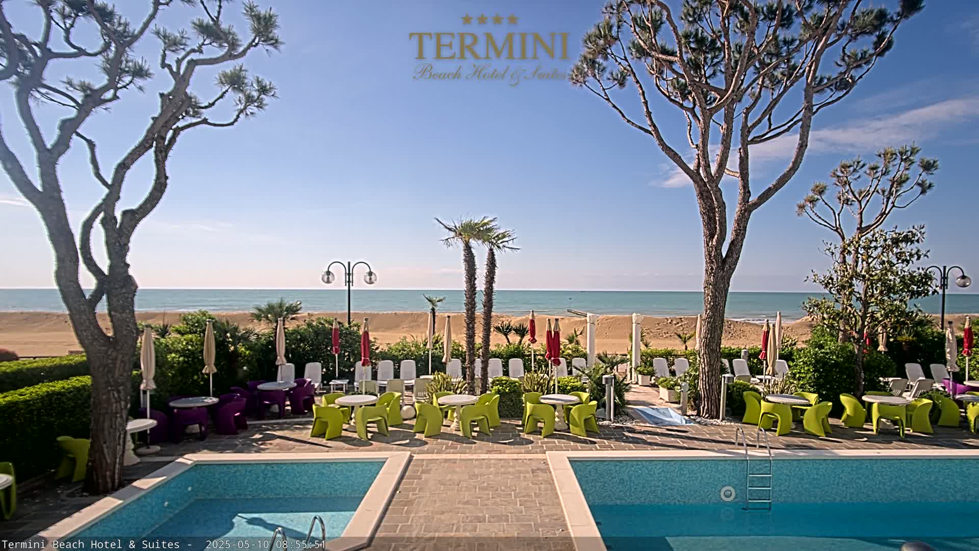 A hotel pool area with bright green and purple chairs and tables sits between two pools overlooking a sandy beach and calm ocean under a clear blue sky.