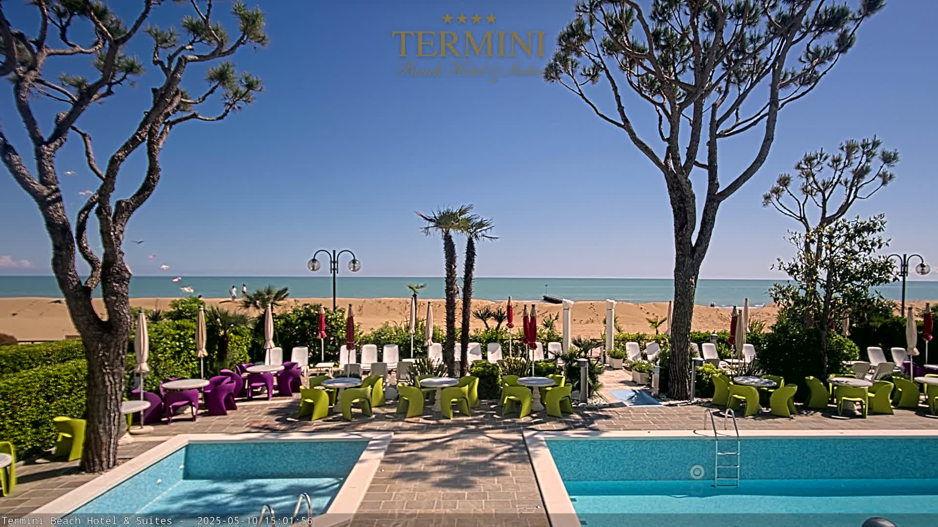 A patio with two pools and colorful outdoor furniture overlooks a sandy beach and calm ocean under a clear blue sky.