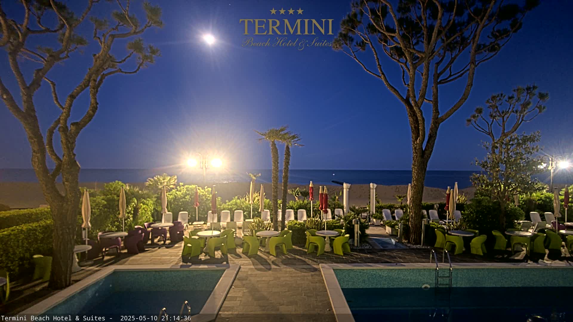 A moonlit beach scene at night shows a hotel patio with two pools, outdoor furniture, and palm trees overlooking a calm ocean.