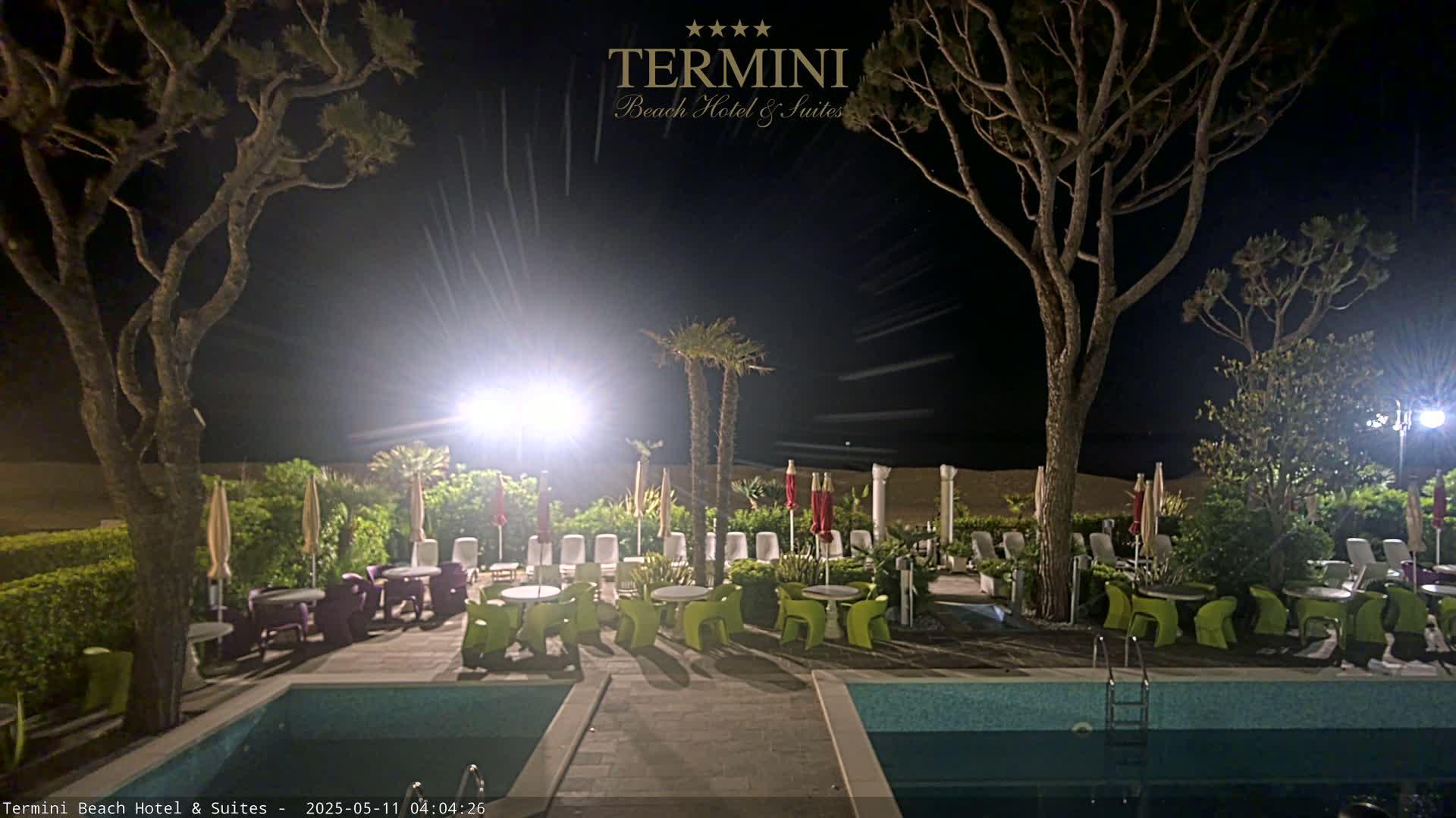 At night, an outdoor patio area with a swimming pool is illuminated, featuring lounge chairs under umbrellas, small tables with chairs, and tall trees flanking the scene.