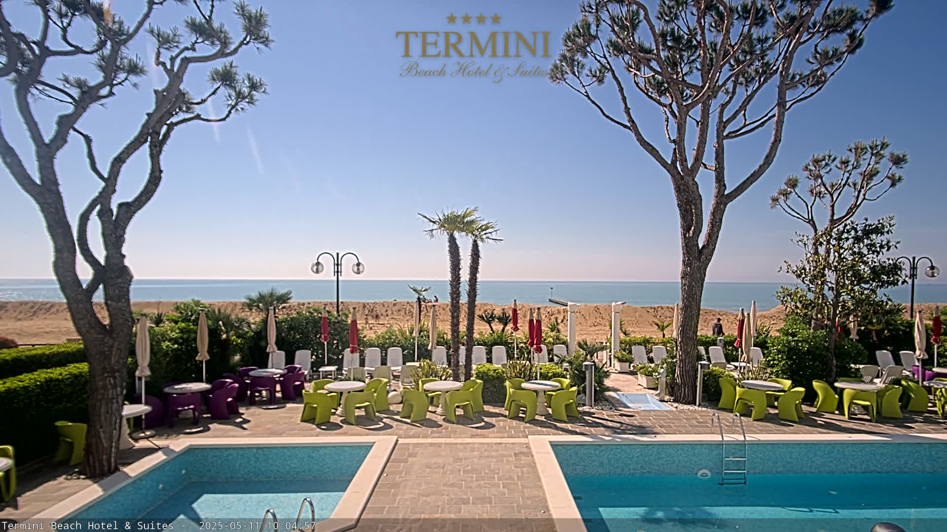 A resort pool area with colorful patio furniture overlooks a sandy beach and calm ocean under a clear blue sky.