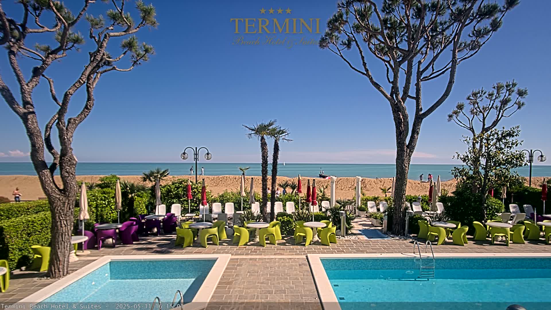 A hotel pool area with colorful furniture overlooks a sandy beach and calm ocean under a clear blue sky.