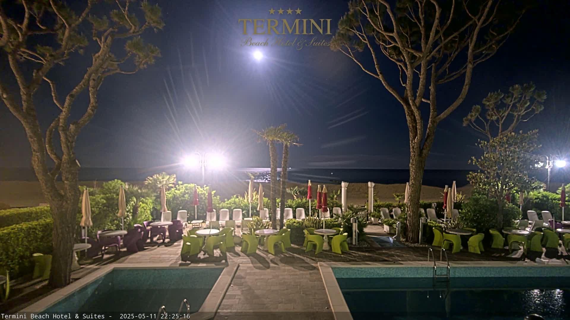 A moonlit night scene shows a hotel pool area with patio furniture, palm trees, and a sandy beach beyond.