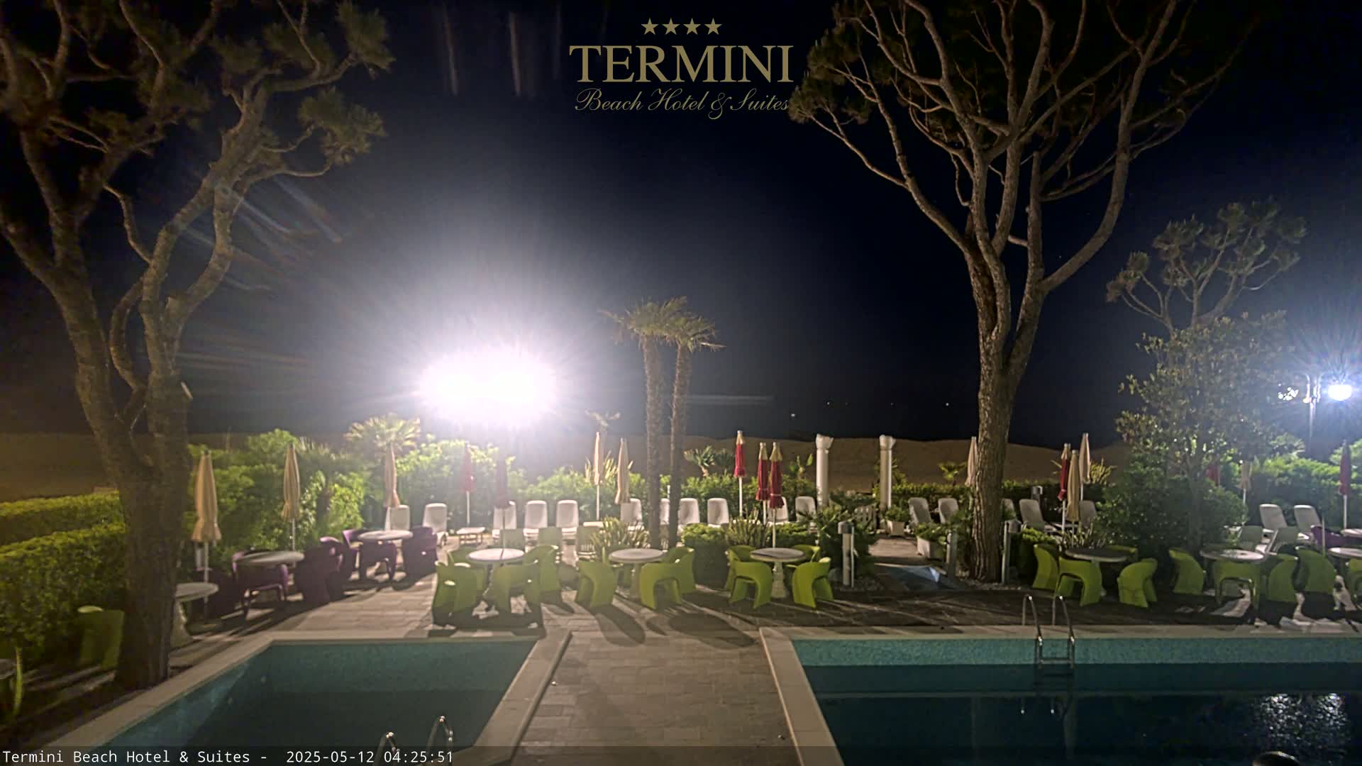 At night, an outdoor area with a swimming pool, patio furniture under umbrellas, and palm trees is illuminated, adjacent to a sandy beach.