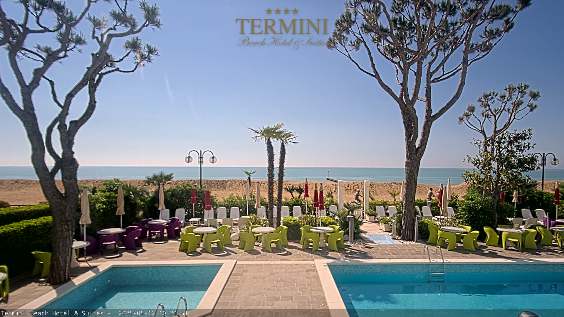 A sunny outdoor scene shows a hotel pool area with colorful chairs and tables, overlooking a sandy beach and calm ocean.