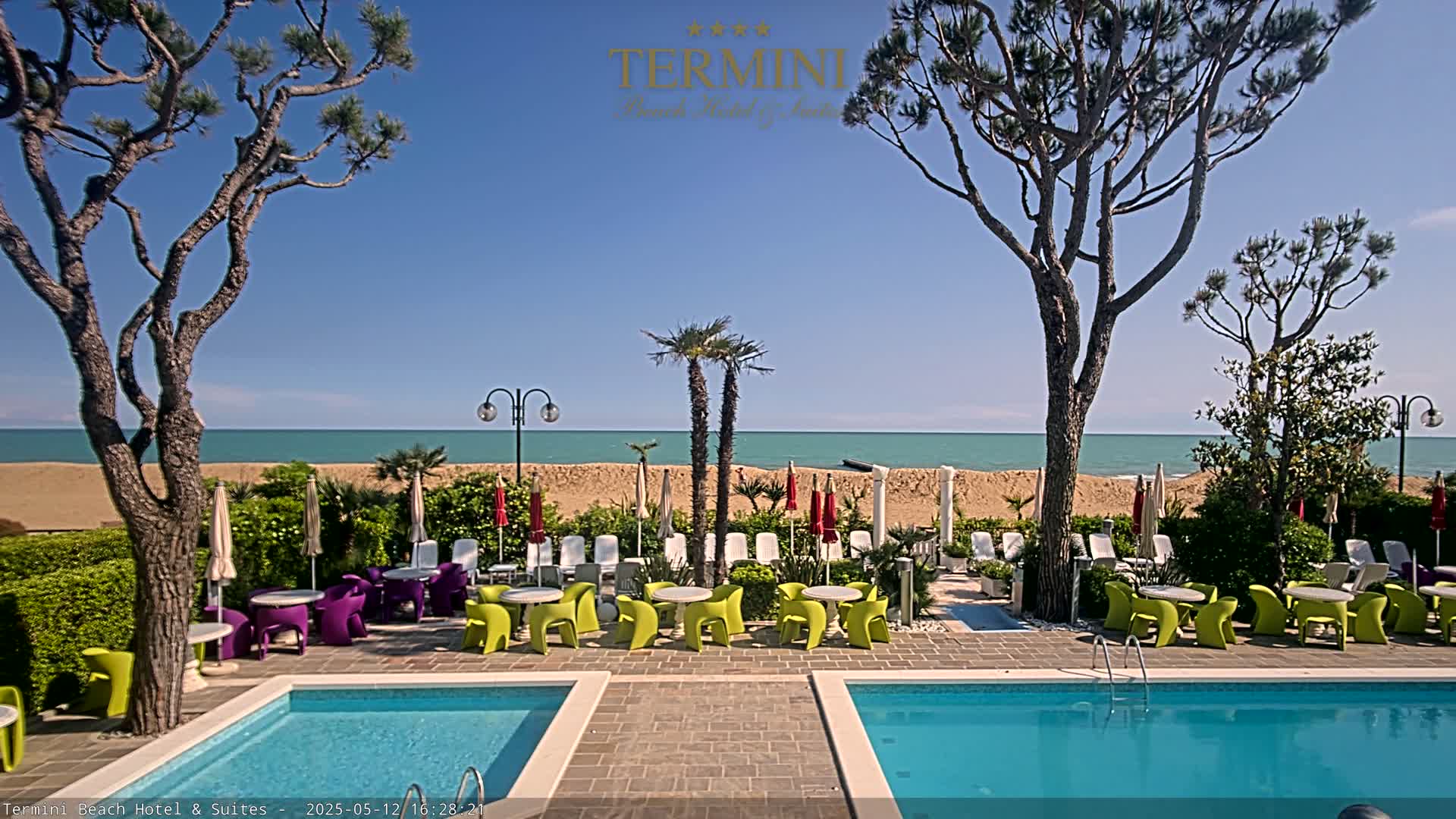 A resort pool area with colorful chairs and umbrellas surrounding it, overlooking a sandy beach and calm ocean under a clear blue sky.