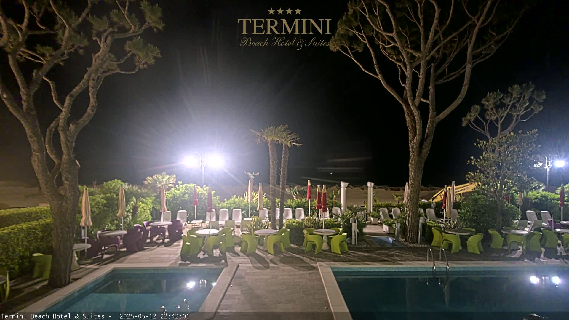 At night, an outdoor patio area with two swimming pools, patio furniture, and tall trees is illuminated by bright lights, overlooking a sandy beach.