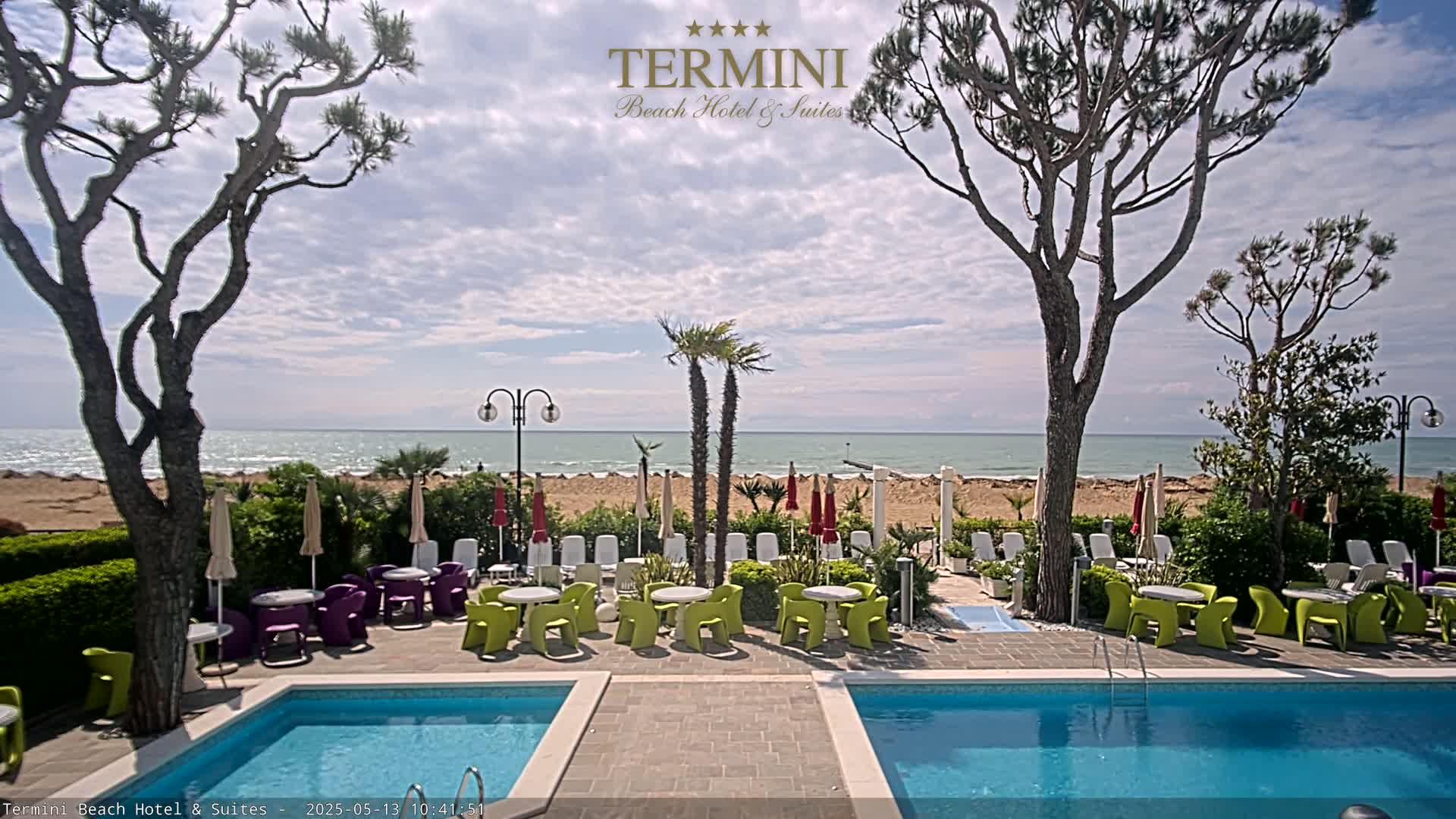 A hotel pool area with patio furniture overlooks a sandy beach and calm ocean under a partly cloudy sky.