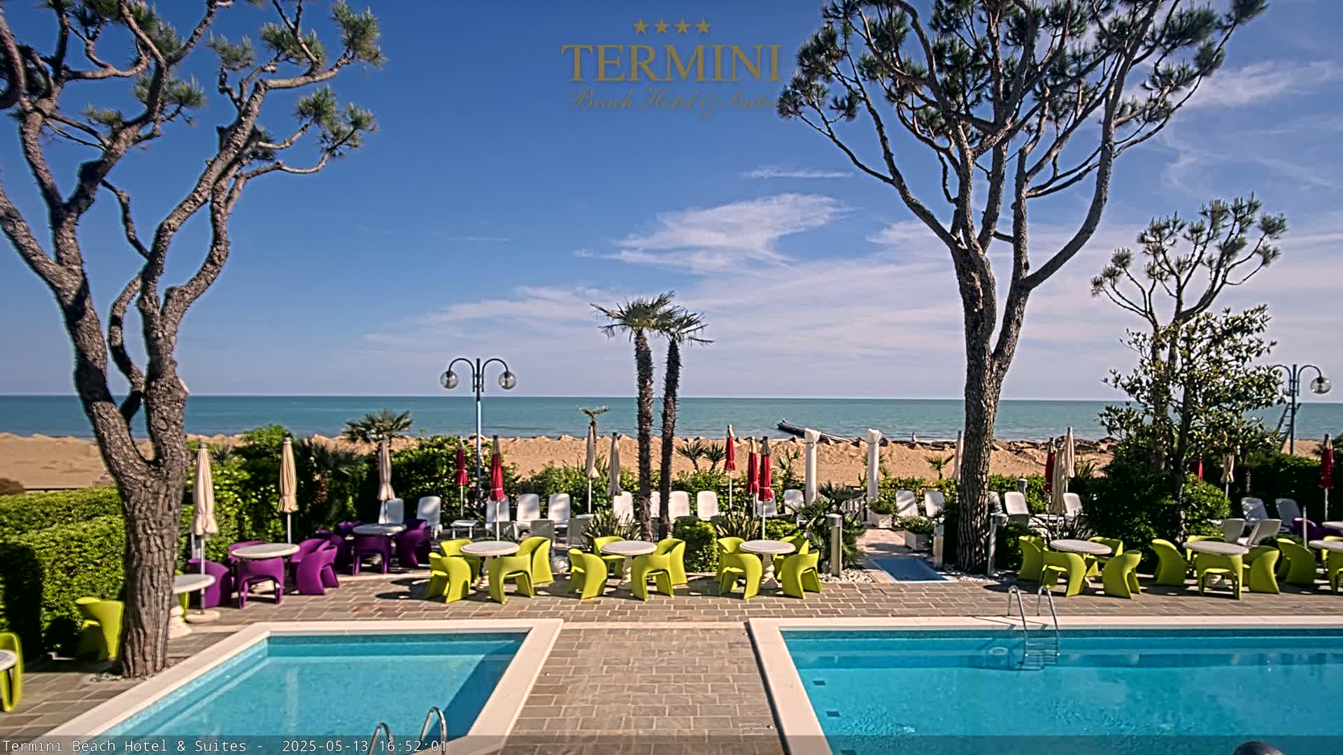 A resort pool area with colorful outdoor furniture overlooks a sandy beach and calm ocean under a sunny sky.