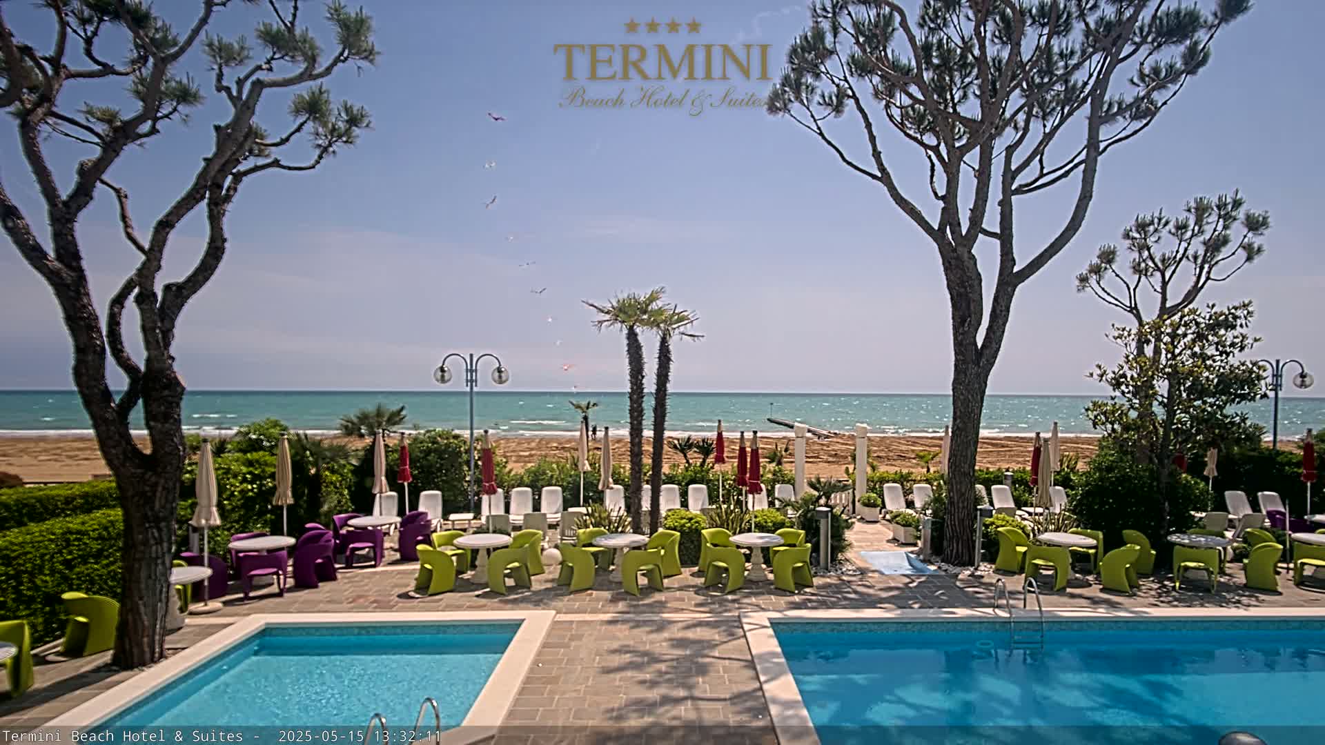 A resort pool area with two pools and outdoor seating overlooks a sandy beach and a calm ocean under a sunny sky.