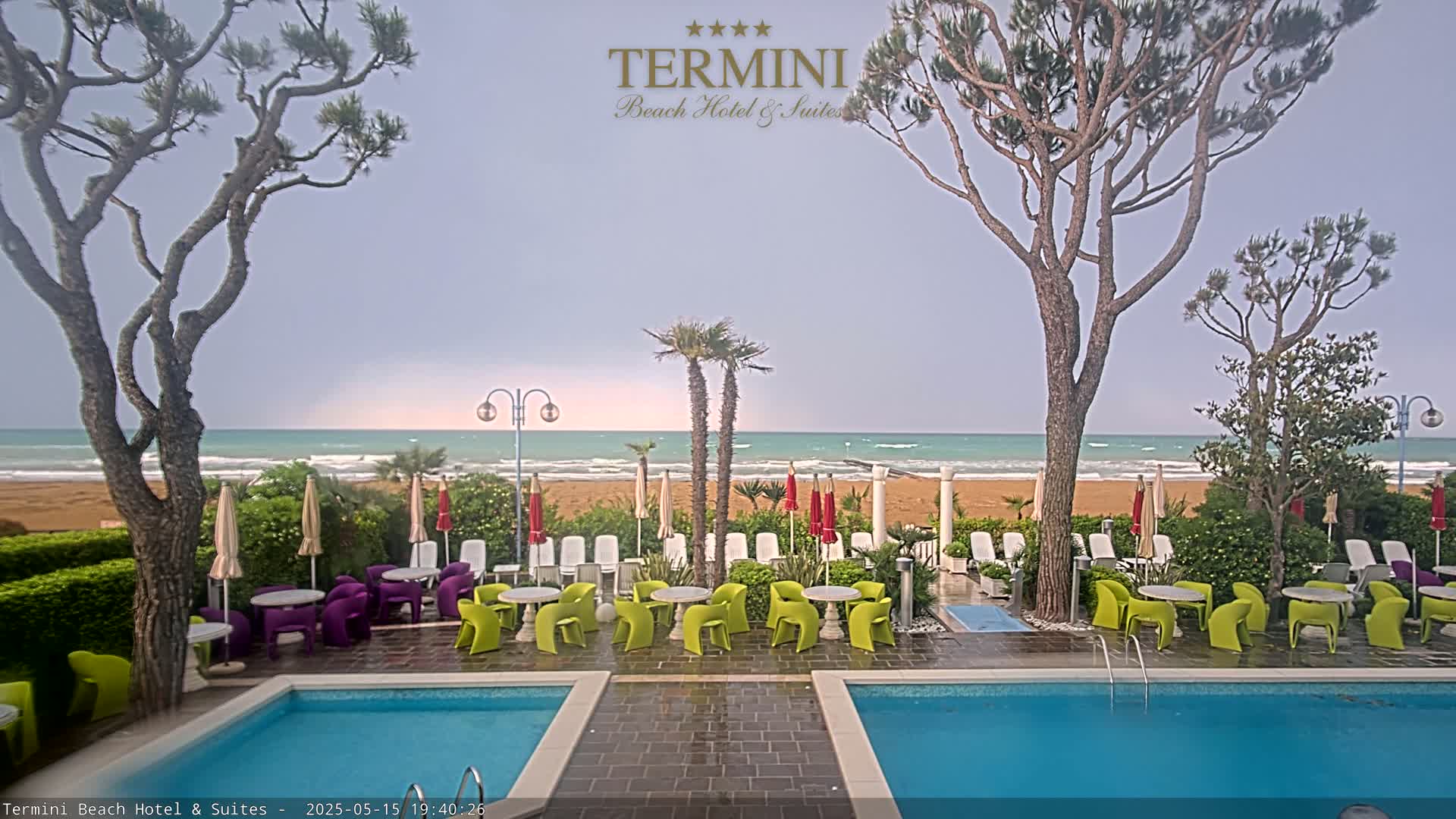 A beachfront hotel pool area with patio furniture, palm trees, and a partly cloudy sky over a wavy ocean.