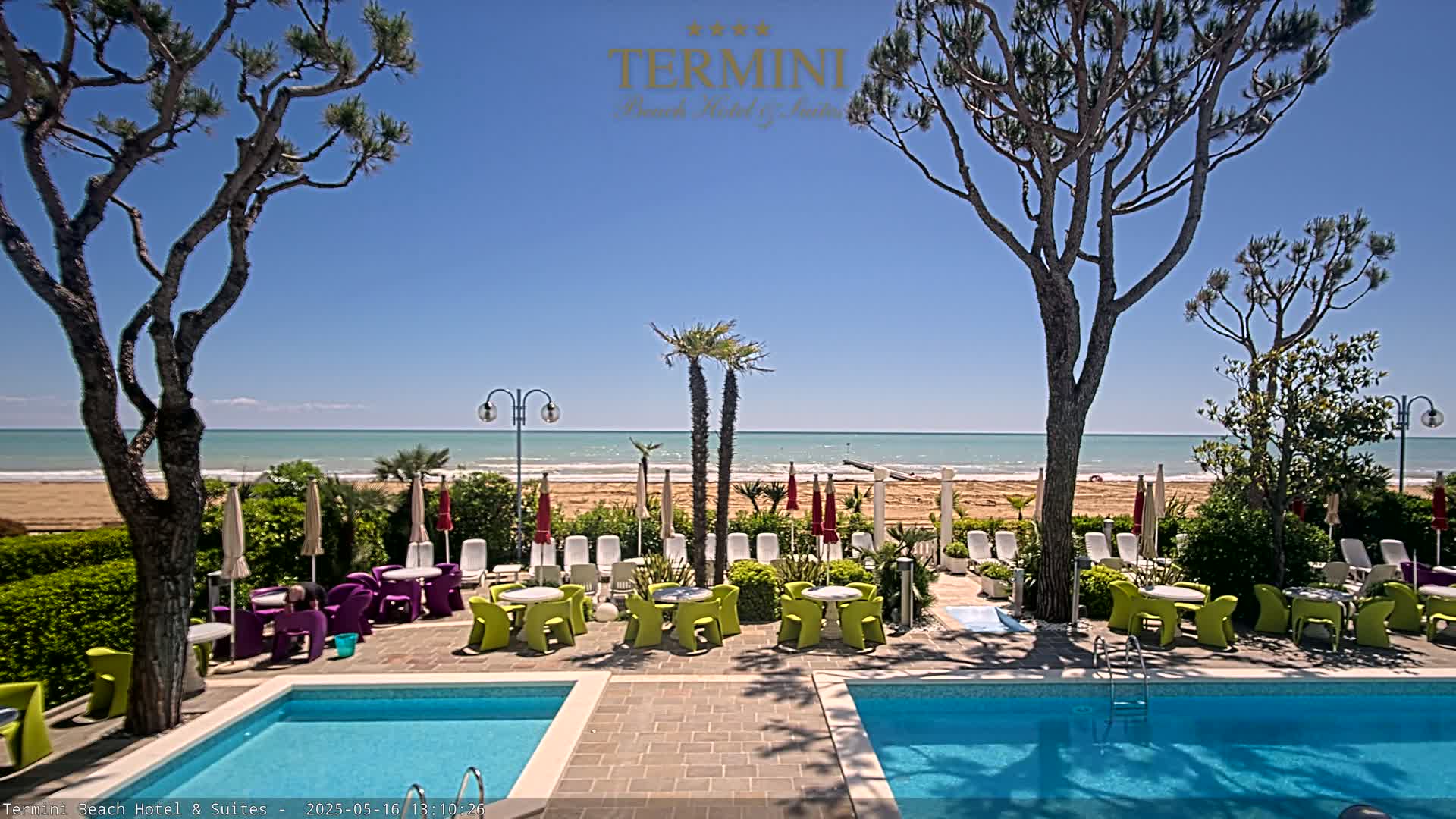 A sunny day at a beachfront resort features two swimming pools surrounded by patio furniture and palm trees, with a sandy beach and calm ocean in the background.