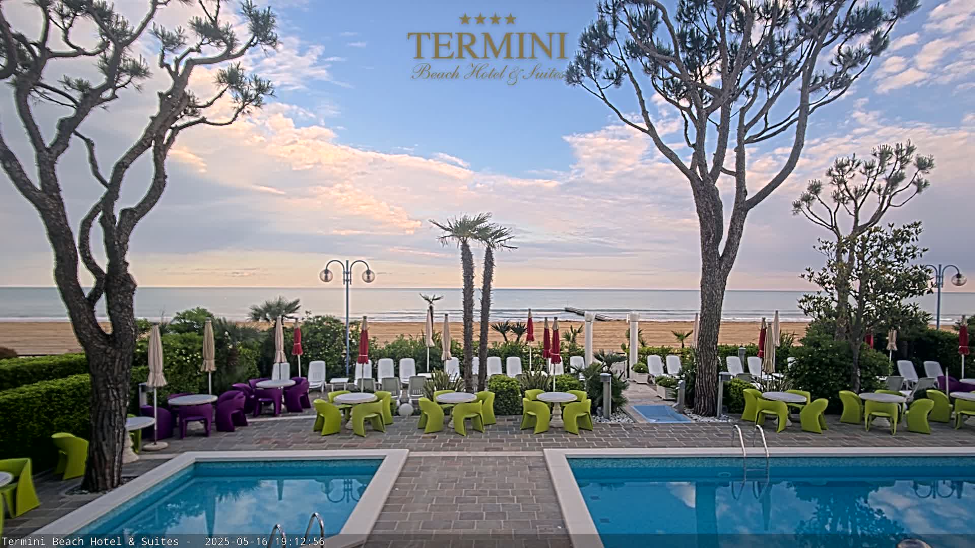 A hotel pool area with two pools overlooks a sandy beach and calm ocean under a partly cloudy sky.
