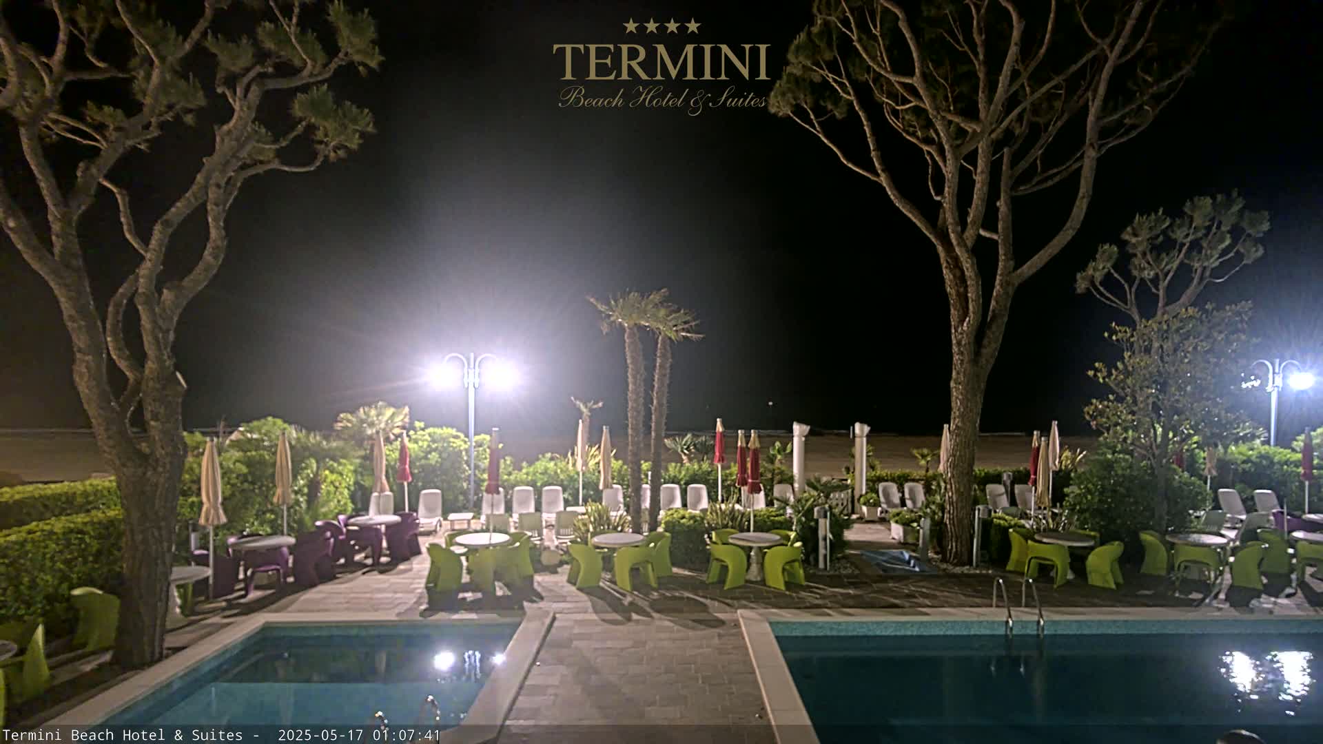 At night, a hotel pool area with lounge chairs, tables, and umbrellas sits near a beach under bright lights, surrounded by trees.