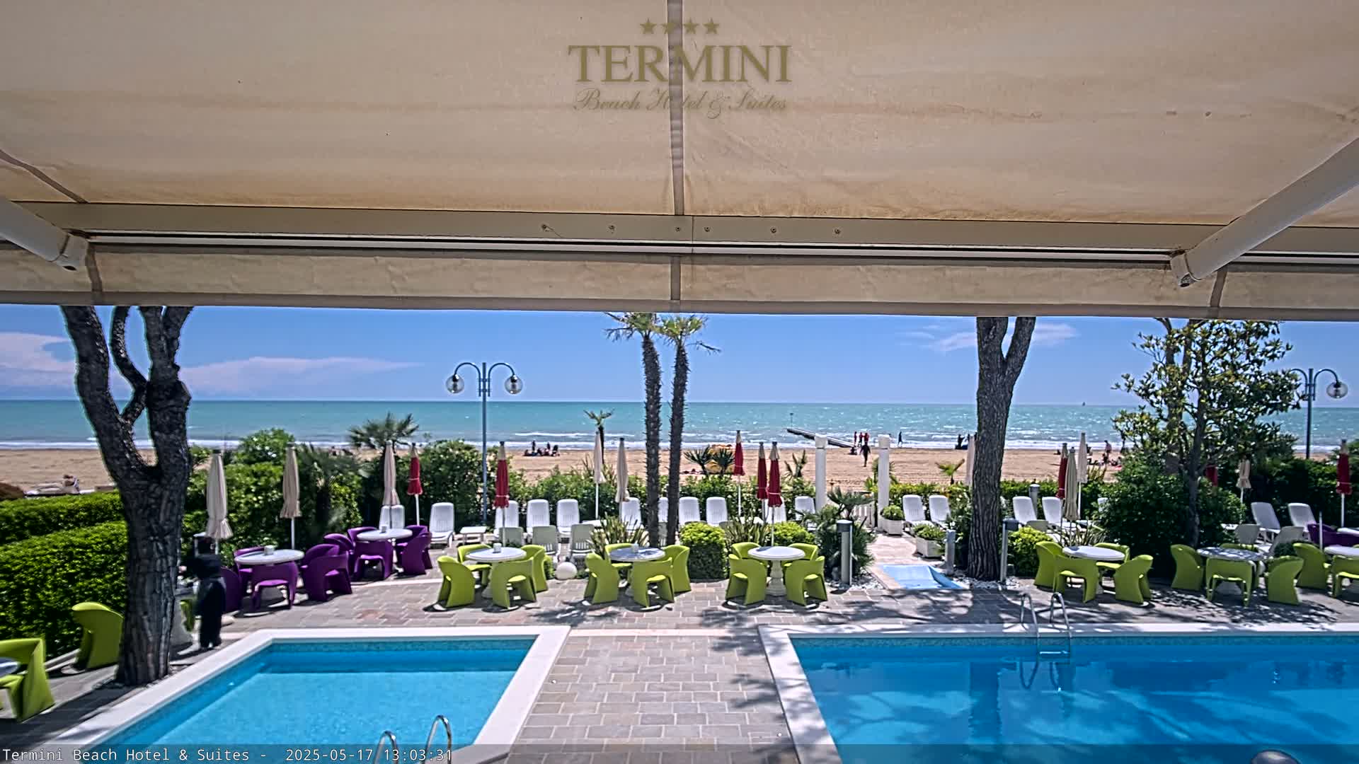 A sunny outdoor scene shows two swimming pools, patio furniture, and a beach with people on it, all viewed from under a patio awning.