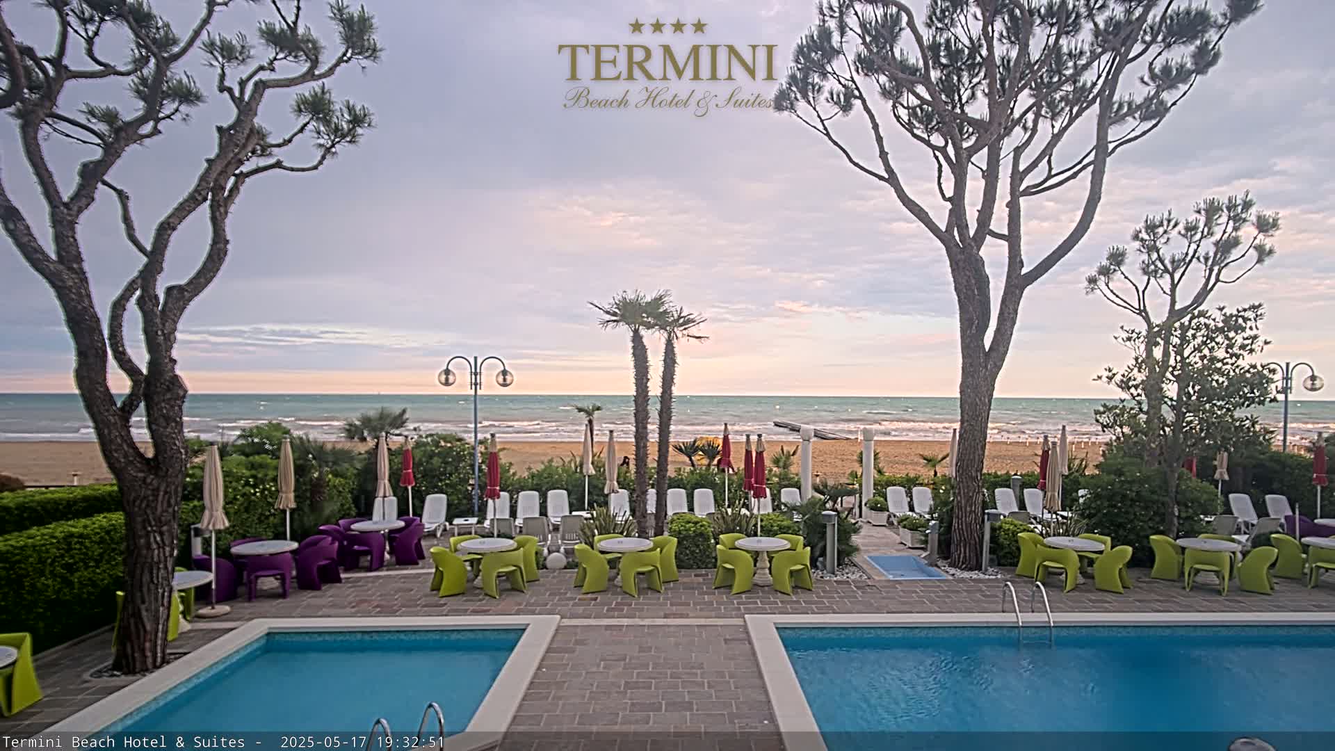 A hotel pool area with patio furniture overlooks a calm ocean beach under a partly cloudy sky.