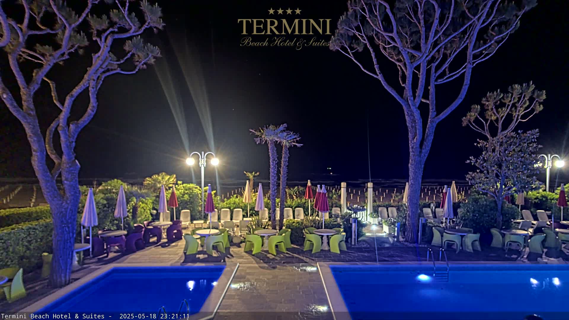 A nighttime view of a resort pool area with lounge chairs, tables, and umbrellas surrounding two illuminated pools, under the shade of trees and near a beach.