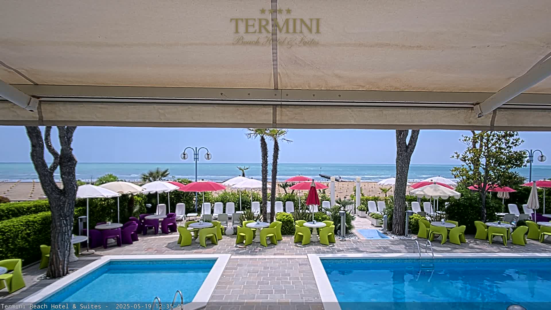 A sunny outdoor scene shows a hotel pool area with colorful tables and chairs under a retractable awning, overlooking a beach with umbrellas and the ocean beyond.