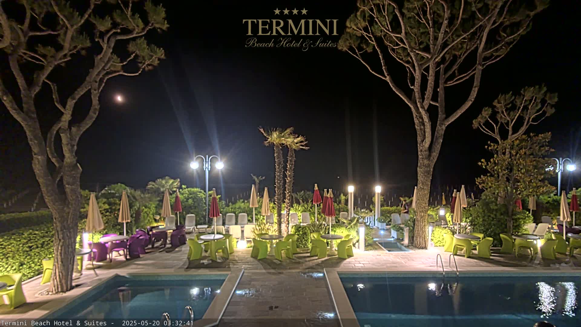 A nighttime outdoor scene shows a pool area with lounge chairs, tables, and umbrellas surrounding two pools under the light of several lamps and a moonlit sky.