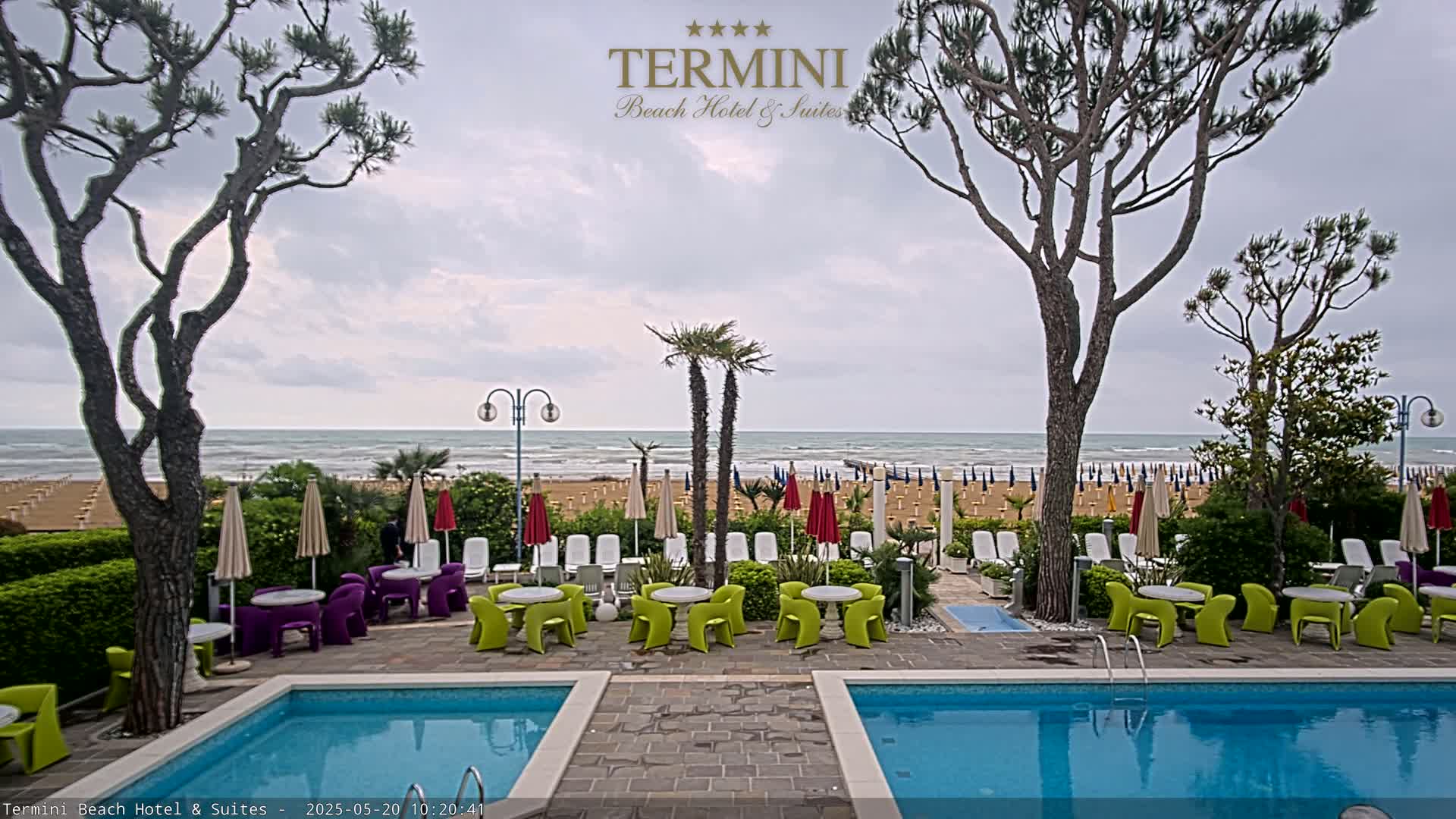 An outdoor pool area with lounge chairs and tables overlooks a beach with numerous umbrellas under an overcast sky.